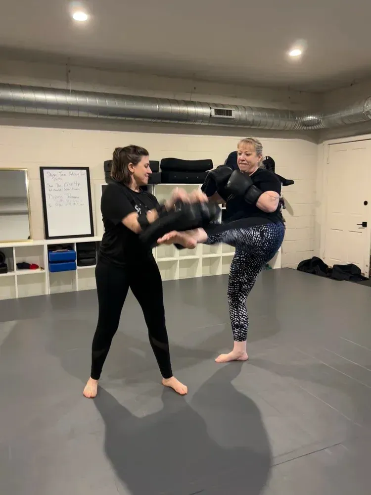 Two people sparring in a martial arts studio, one delivering a kick on a gray mat.