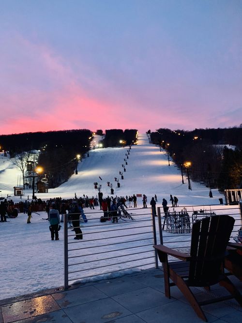 Ski slope at dusk with scattered skiers, chairlift, and pink-blue sky over snowy hills