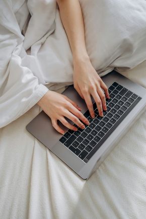 Hands typing on a laptop on a white bed with rumpled bedding