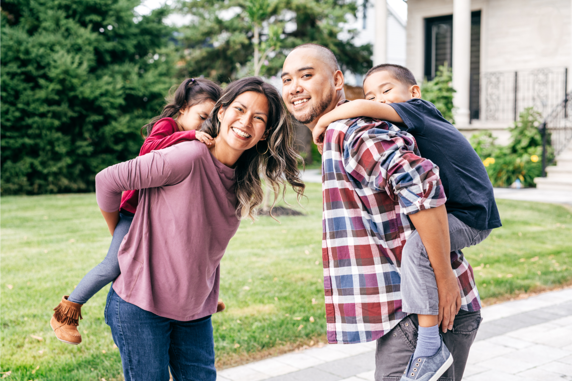 Family with two children, smiling and giving piggyback rides outside a house.