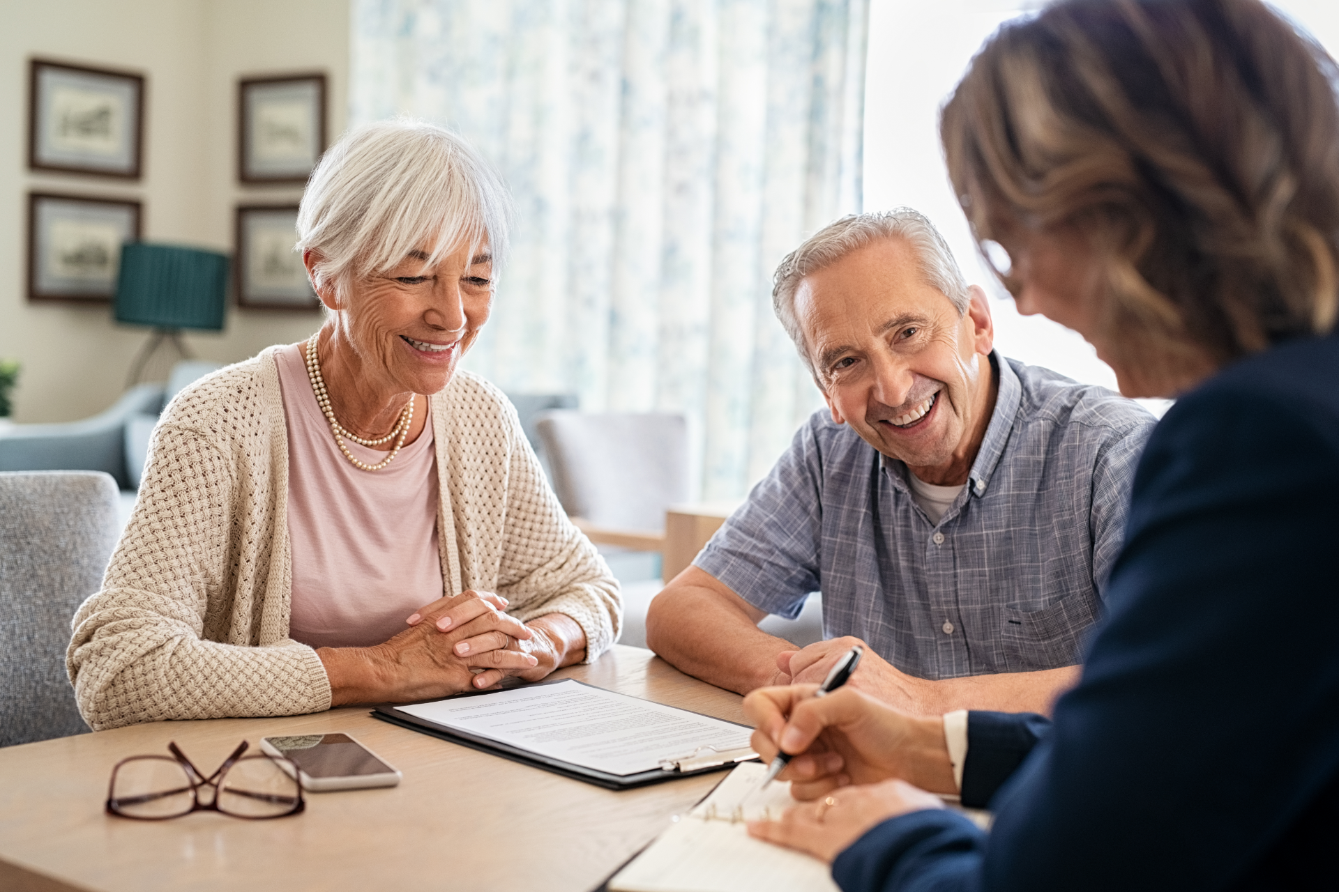 An elderly couple smiling as a person in a suit writes on a document at a table.