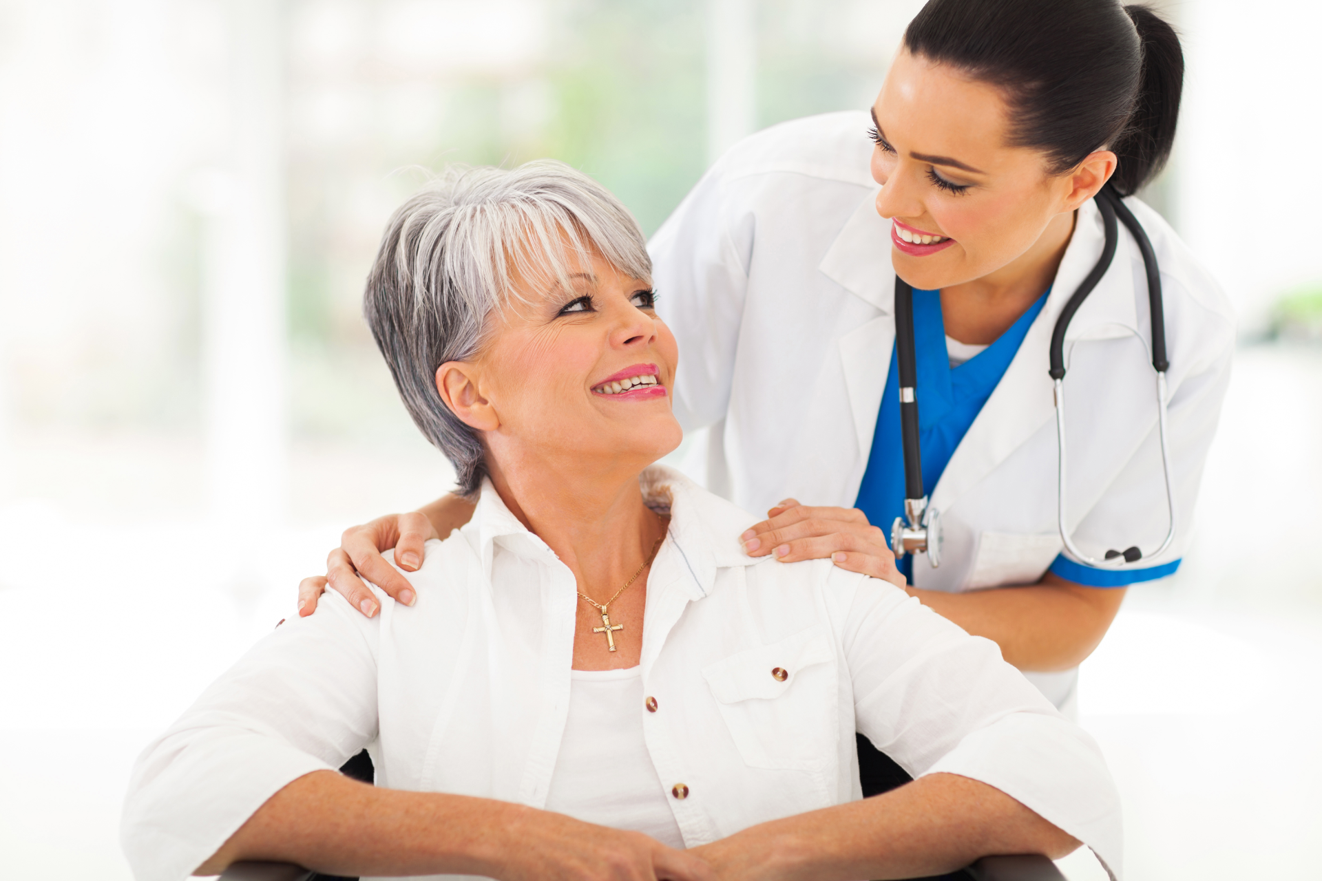 Smiling nurse with hands on shoulders of elderly woman in wheelchair.