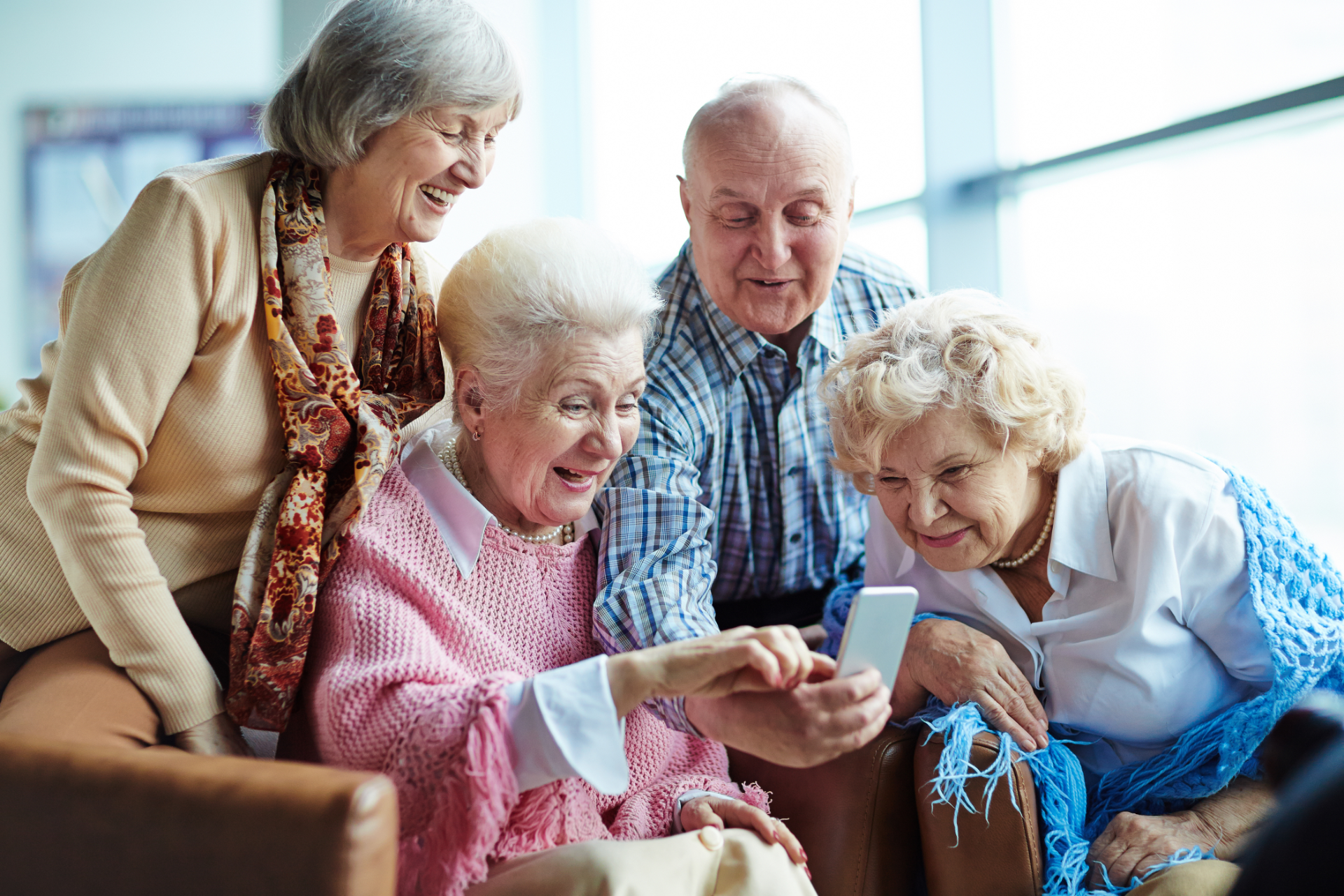 Four older adults smiling while looking at a smartphone; indoors, natural light.