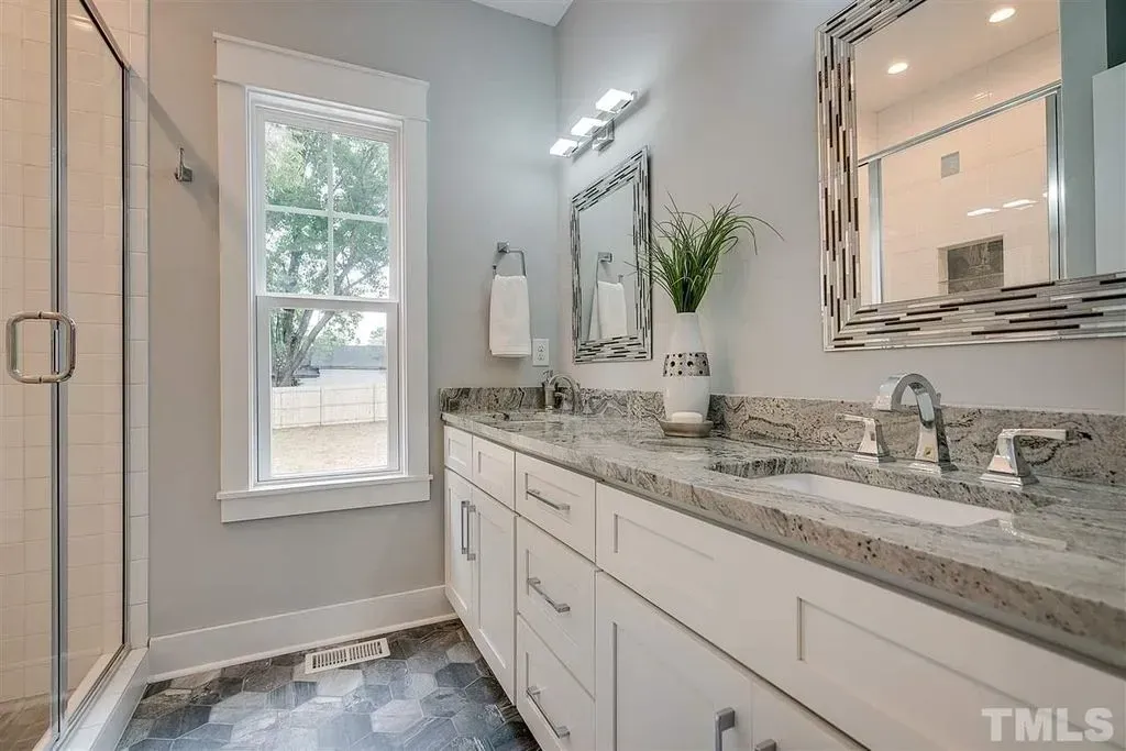 Bright modern bathroom with double vanity, granite countertop, large mirror, and shower stall