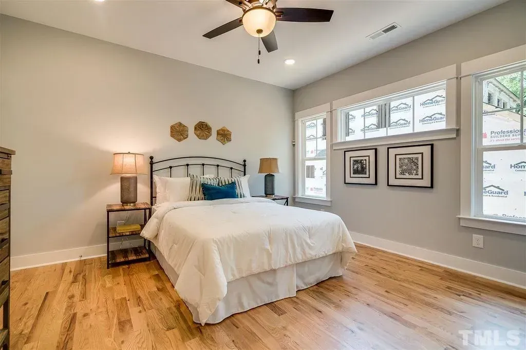 Bright bedroom with white bed, wood floors, ceiling fan, and large windows with framed art