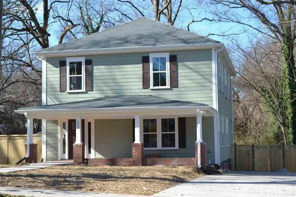 Two-story light green house with a front porch, brick accents, and a driveway in a wooded yard.