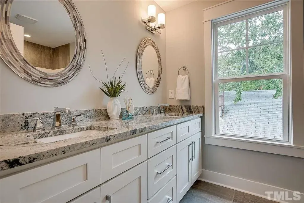 Bright bathroom vanity with granite countertop, round mirrors, wall sconce, and large window.