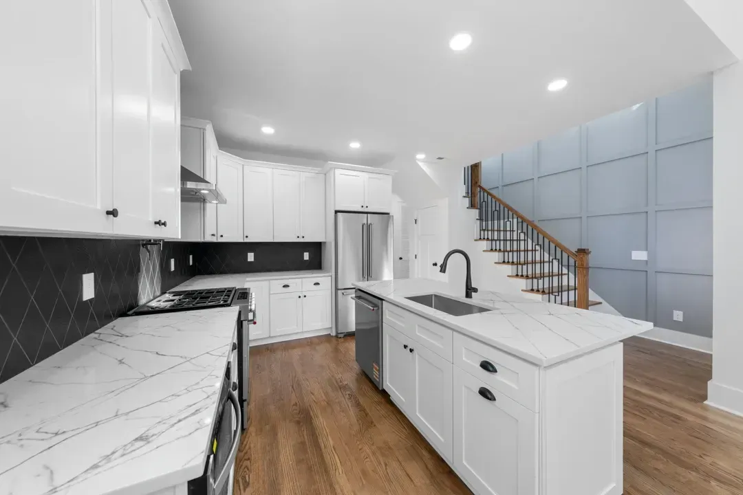 Bright modern kitchen with white cabinets, marble island, black backsplash, wood floors, and staircase
