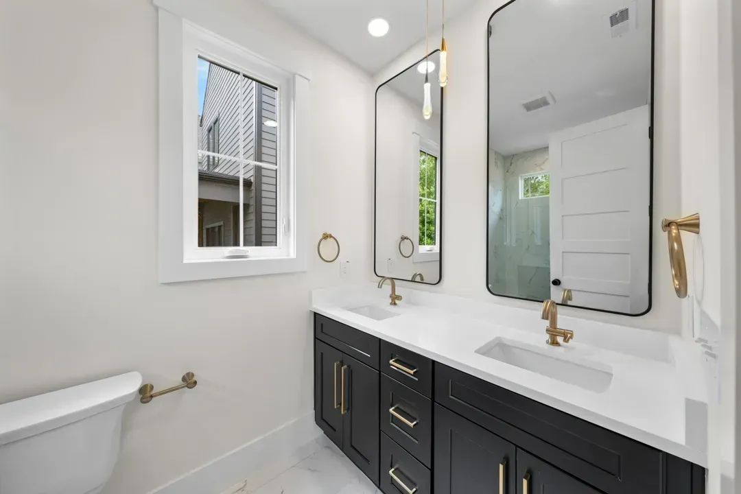 Bright bathroom with double vanity, large mirror, white walls, and a window beside a shower.