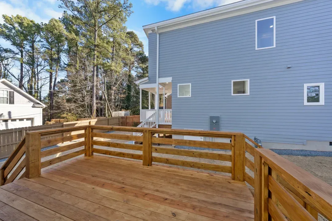 Wooden deck with railing beside a blue house in a sunny backyard.
