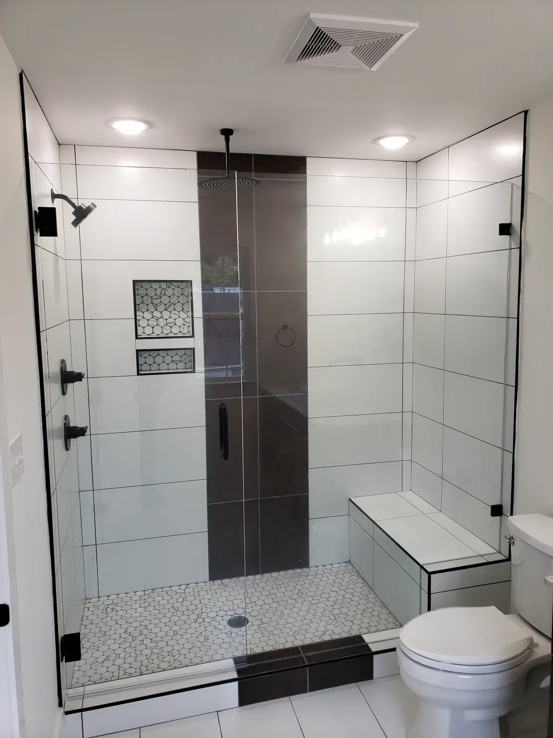 Modern white-tiled shower with glass enclosure, bench, and toilet in a bathroom.