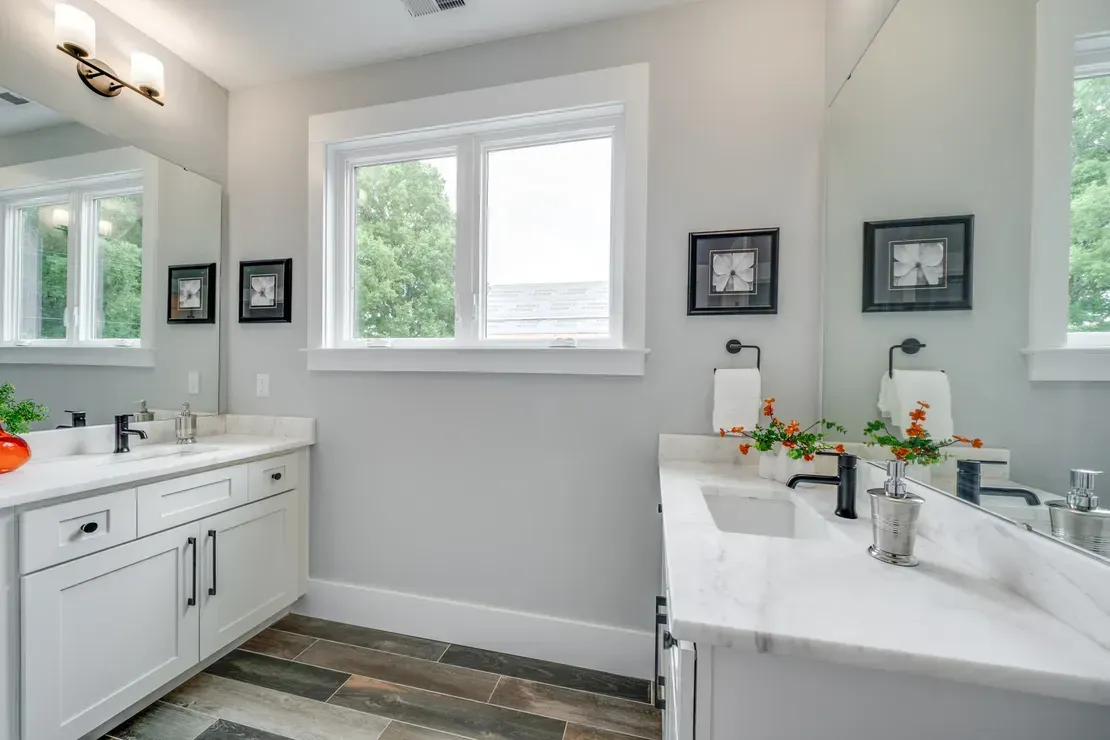 Bright white bathroom with a double vanity, mirror, and window, accented by black fixtures and a gray rug.