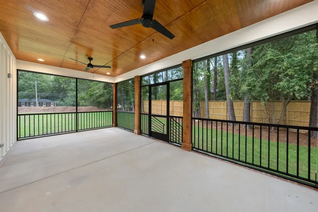 Screened porch with wood ceiling, ceiling fans, and black railing overlooking a grassy yard and trees.