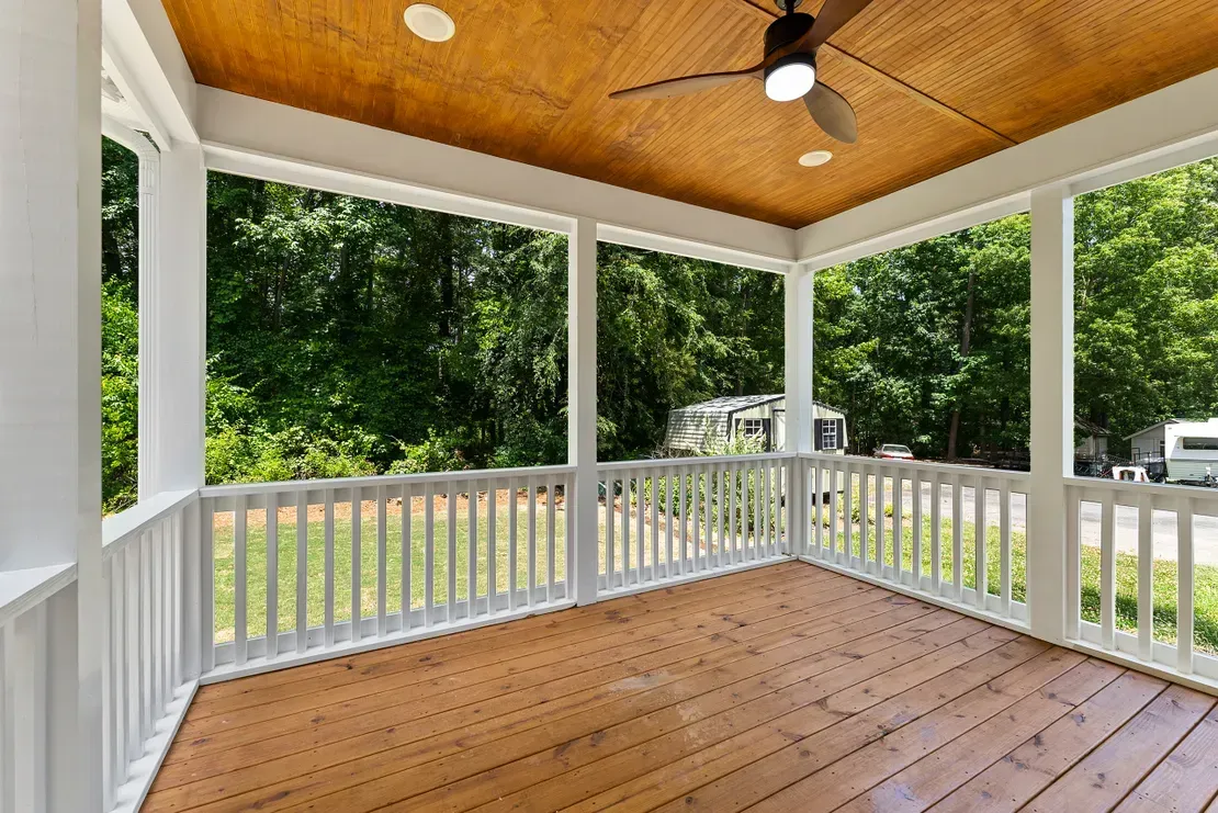 Covered porch with white railings, wood ceiling, and tan floor overlooking trees and parked vehicles.