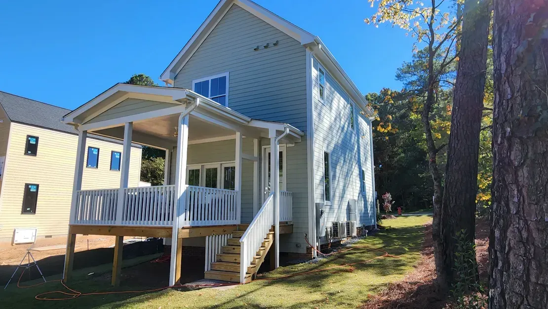 White house with front porch and stairs, beside a brick building and tall trees on a sunny day