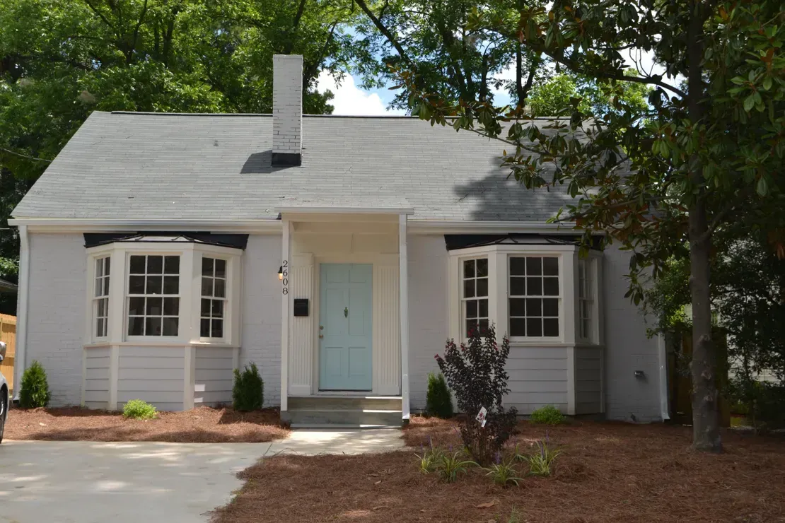 Small gray cottage with a pale blue front door, white trim, and trees shading the yard.