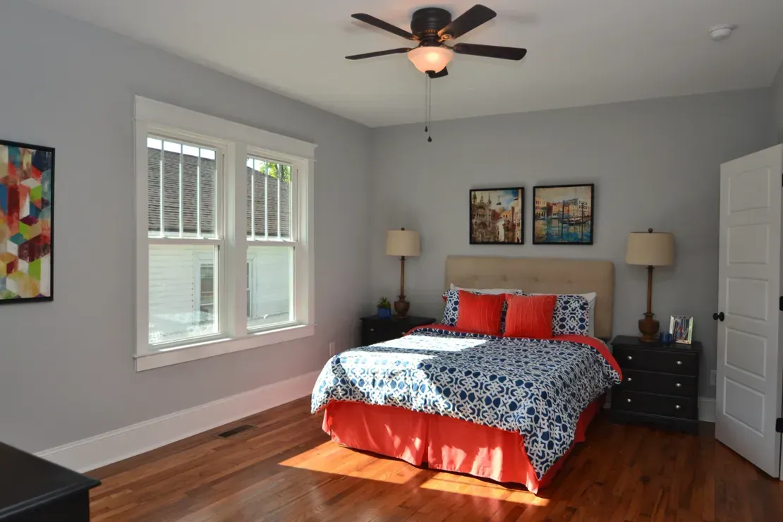 Bright bedroom with a bed, two bedside lamps, ceiling fan, and sunlight on hardwood floors.