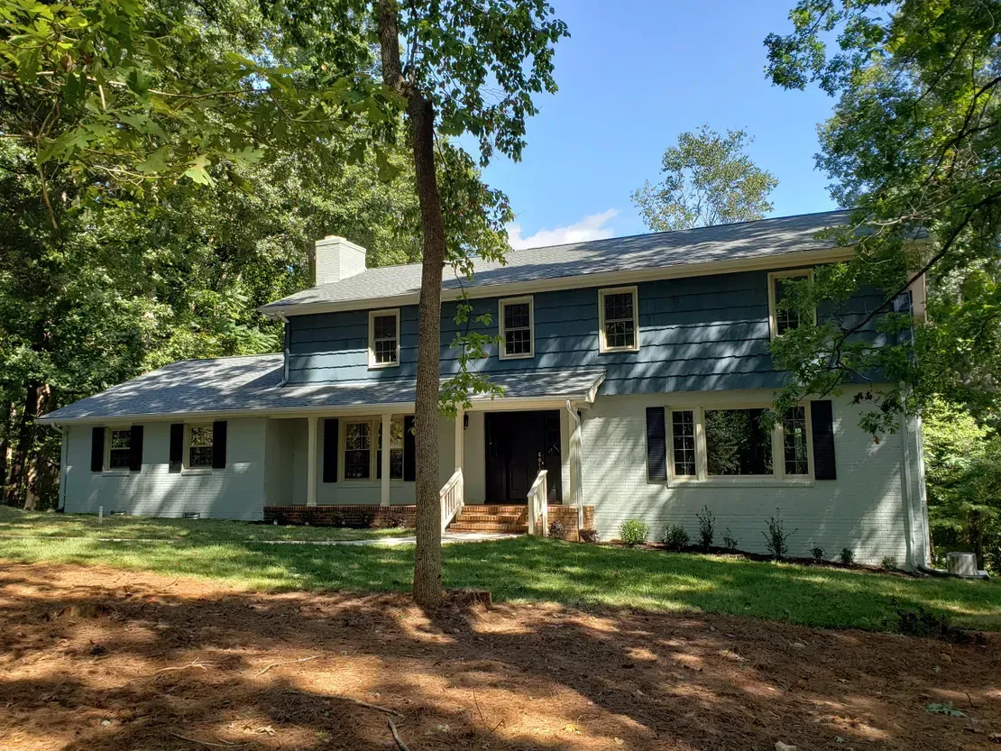 Two-story blue-and-white house surrounded by trees and a wooded yard