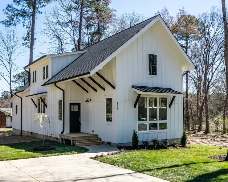 White farmhouse with black trim and porch in a wooded yard under a clear sky