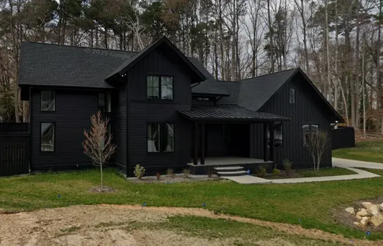 Black modern farmhouse with steep gables and a covered porch on a grassy lot surrounded by trees