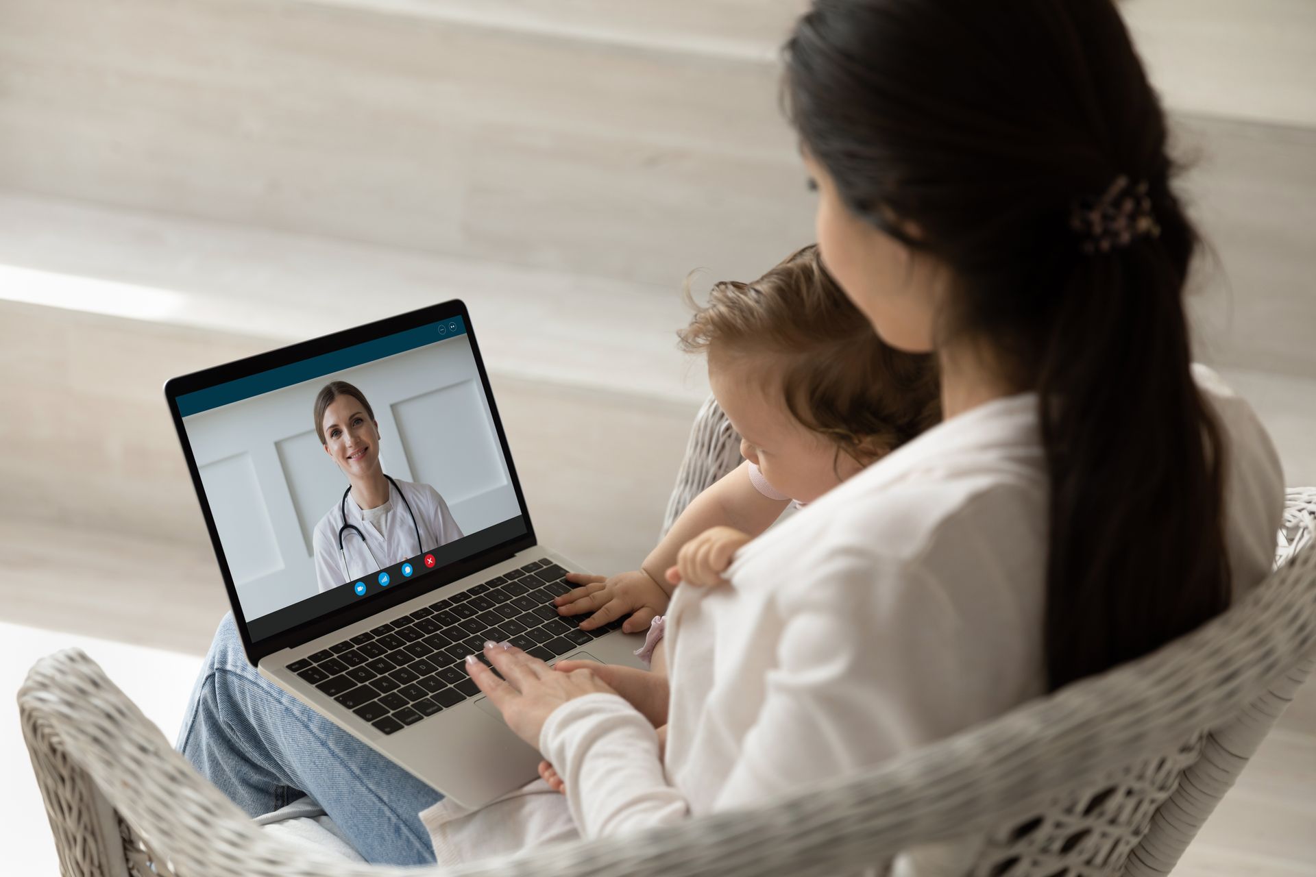 Woman and toddler on a laptop video call with a doctor, likely for a telehealth appointment. They sit in a light-filled room.