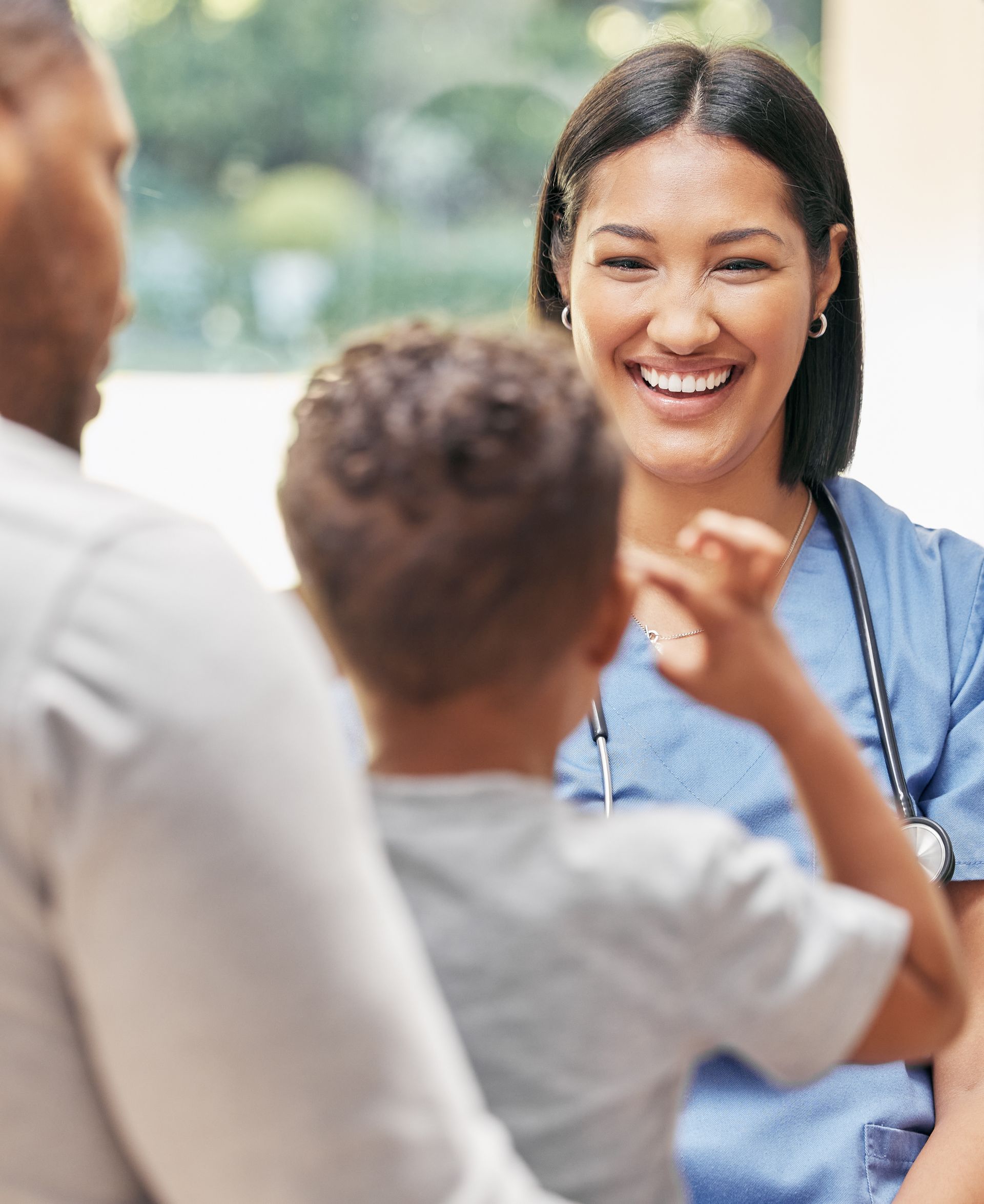 A smiling nurse in scrubs interacts with a young child and a man in a bright setting.