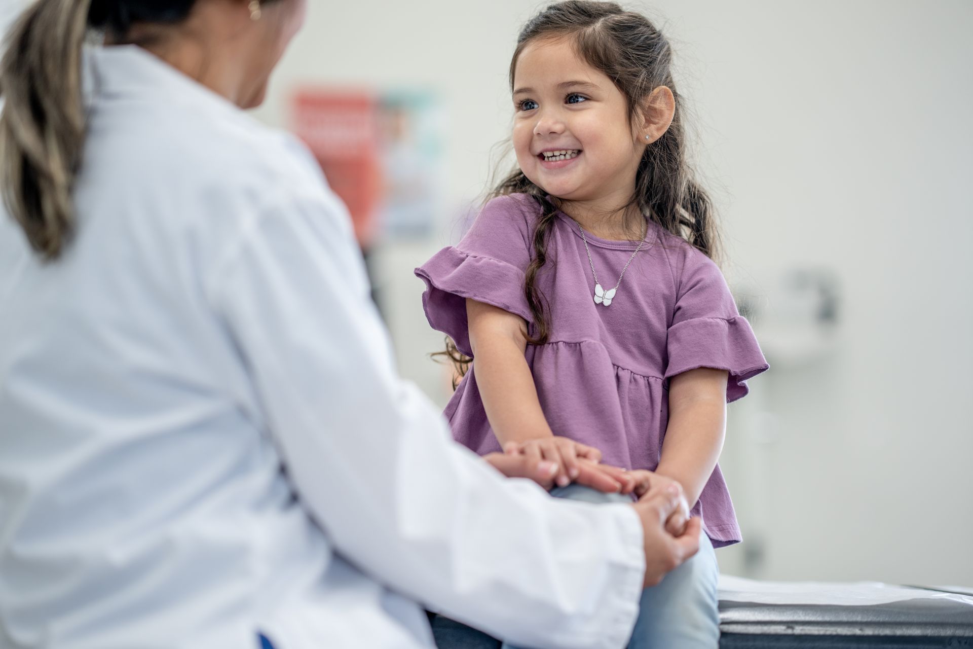 A young girl in a purple shirt smiles at a doctor in a white coat during a medical examination in a brightly lit room.