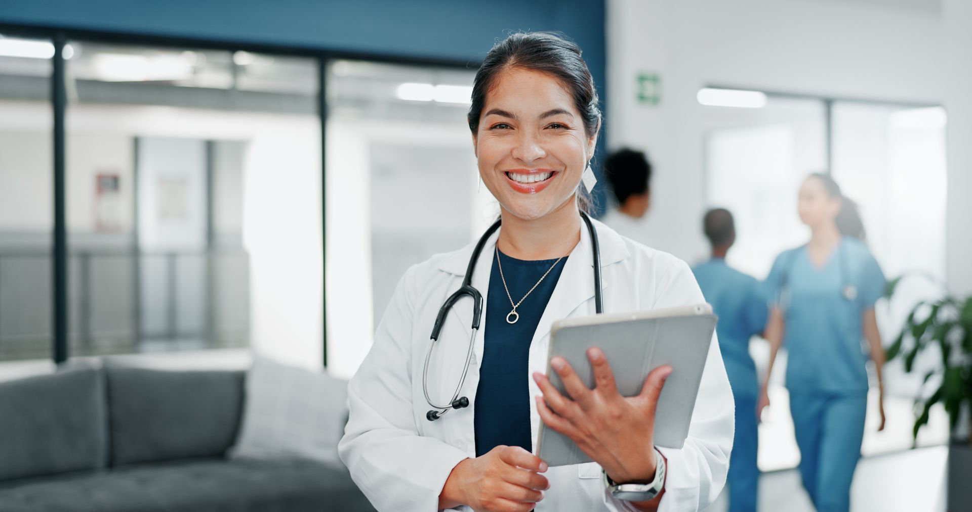 Smiling female doctor in a white coat, holding a tablet, with colleagues and a hospital corridor in the background.