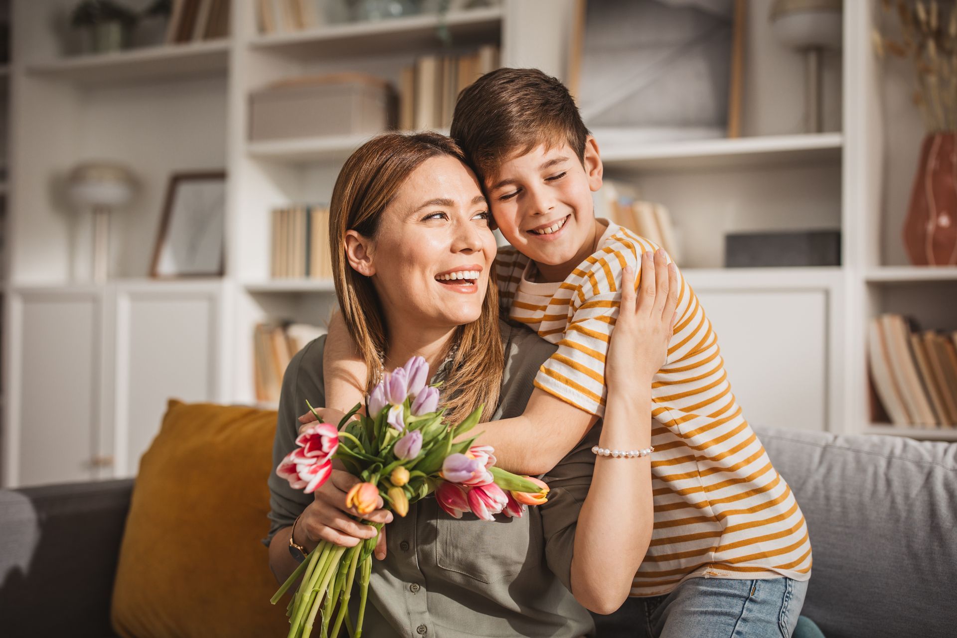 Woman holding flowers smiles as child hugs her on couch.