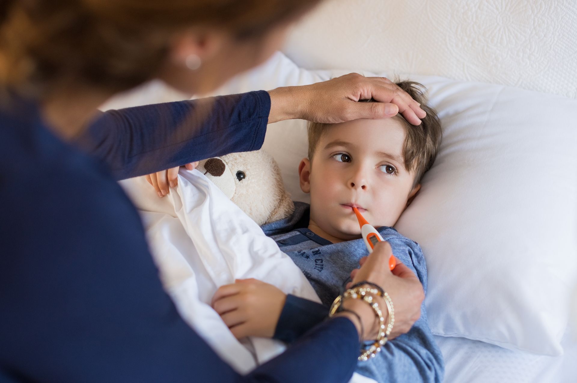A child lies in bed with a thermometer in their mouth while an adult checks their forehead.