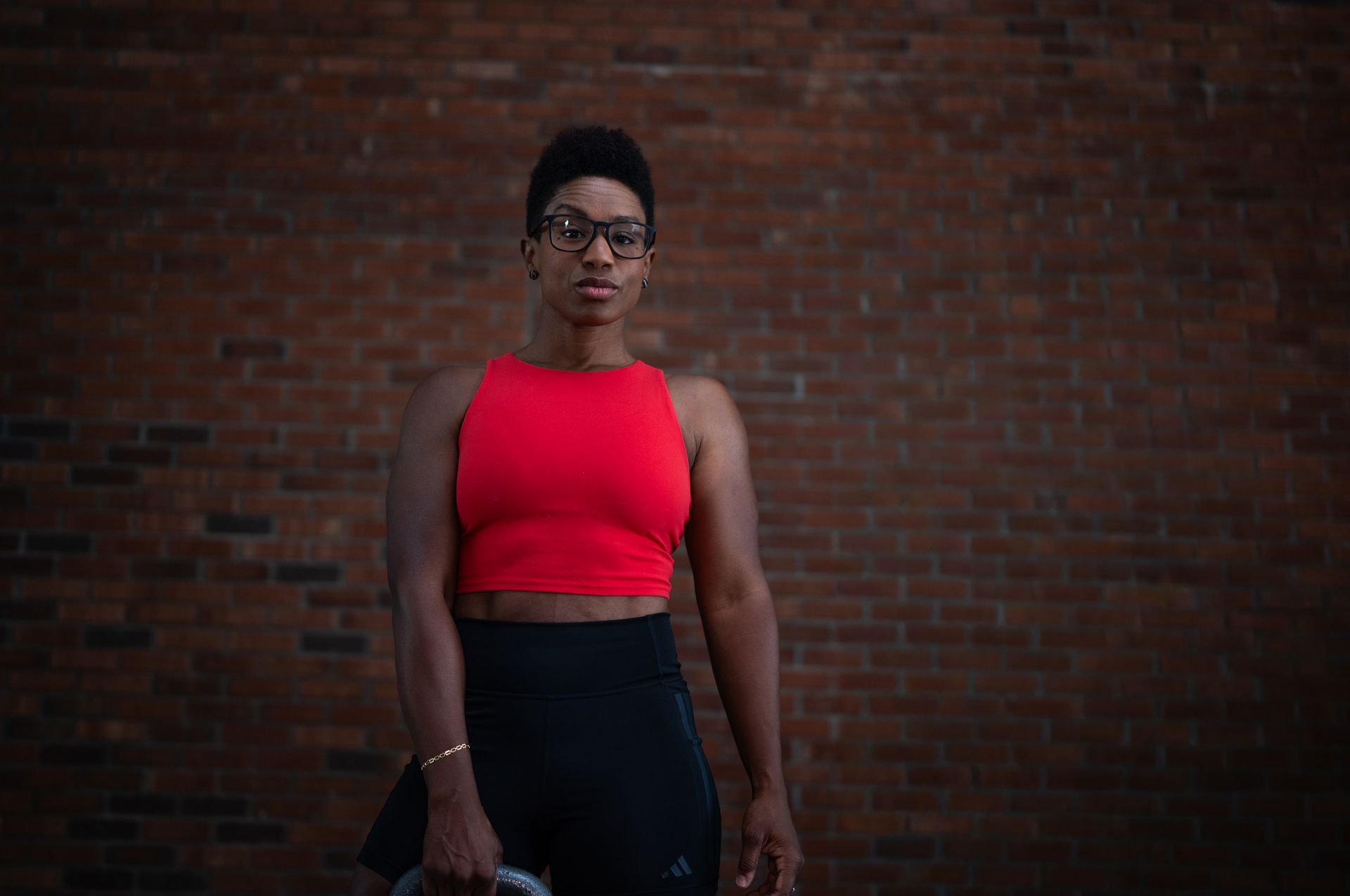 Person in red top and black pants standing against a brick wall, holding a phone.