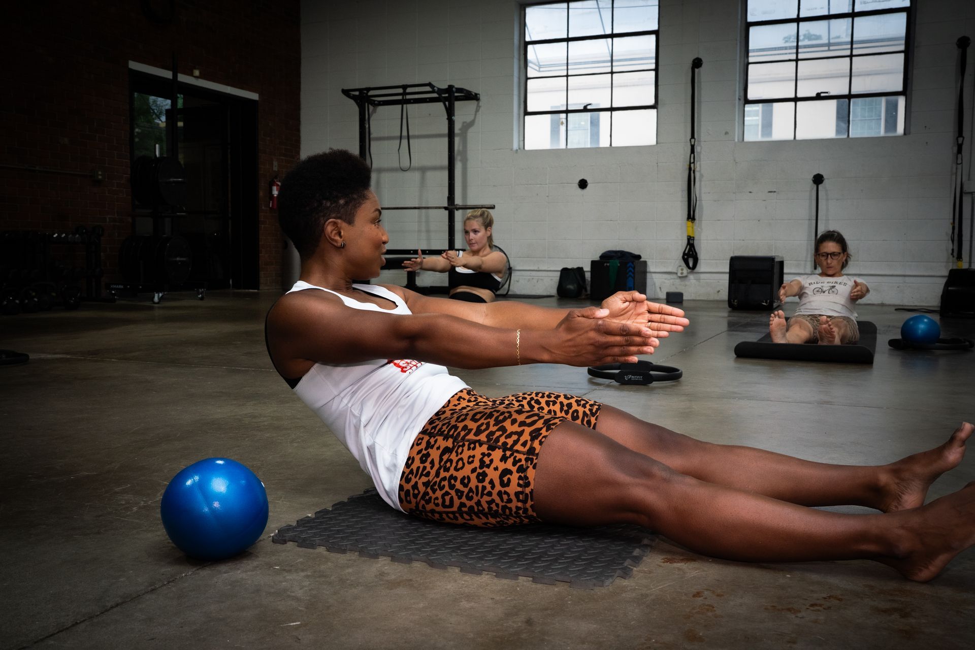 People exercising on yoga mats in a bright studio, with one person doing a seated crunch near a blue ball