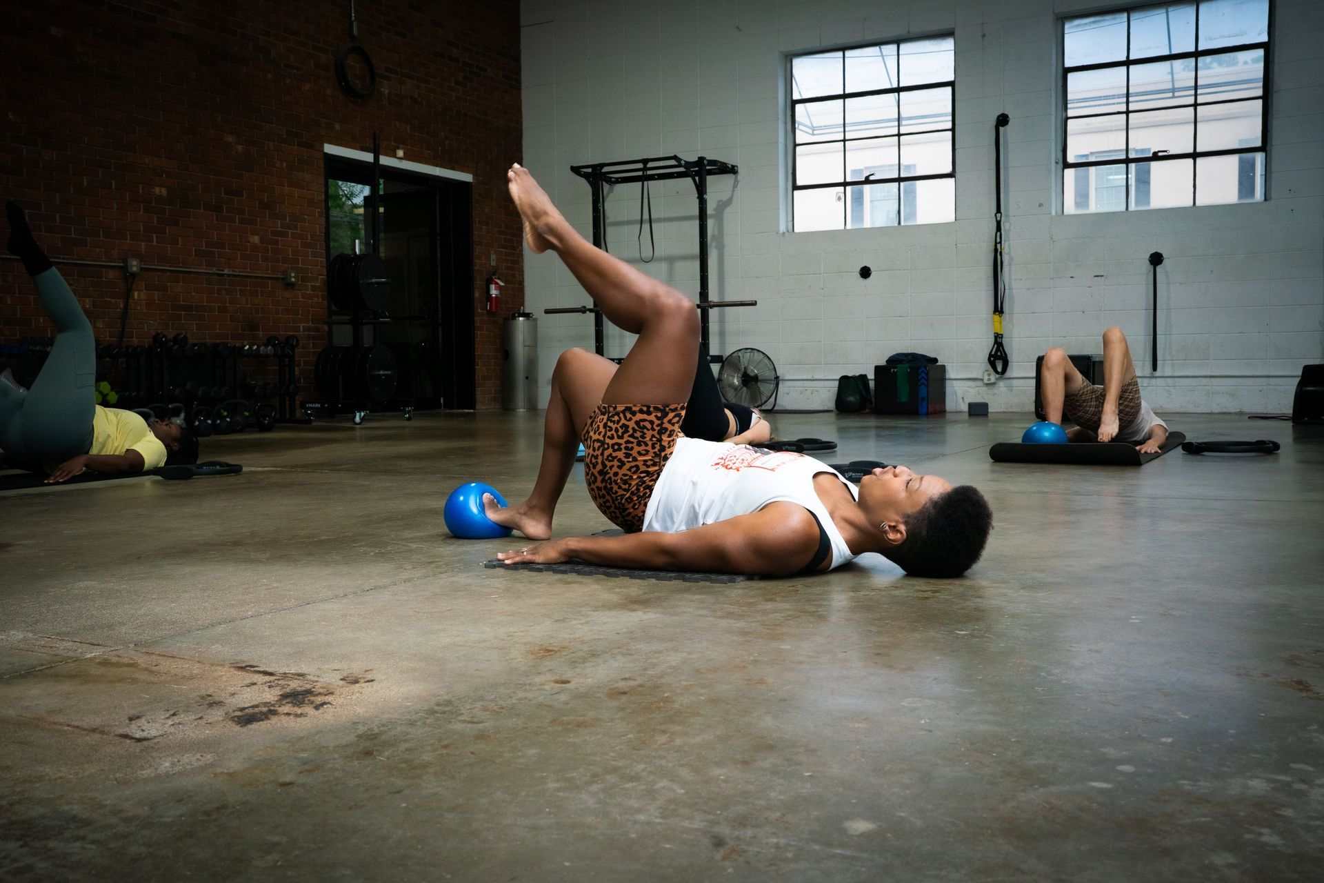 People exercising on mats in a gym, with one person stretching a leg upward in the foreground.