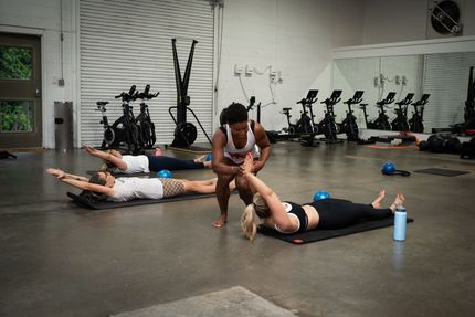 People stretching on gym mats while an instructor helps them in a fitness studio