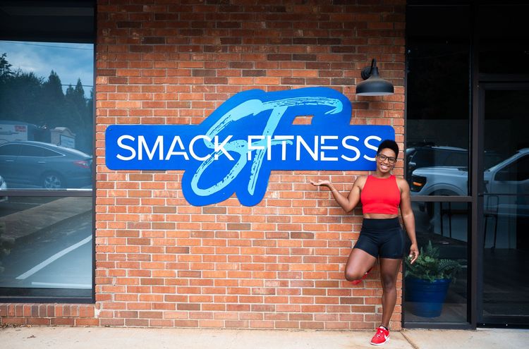 Woman in red athletic wear standing by the SMACK FITNESS sign on a brick wall.