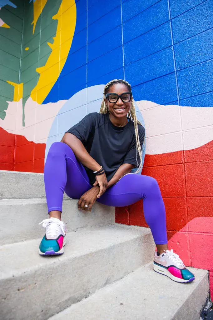 Person sitting on concrete steps in front of a colorful mural, smiling and wearing purple leggings and white sneakers