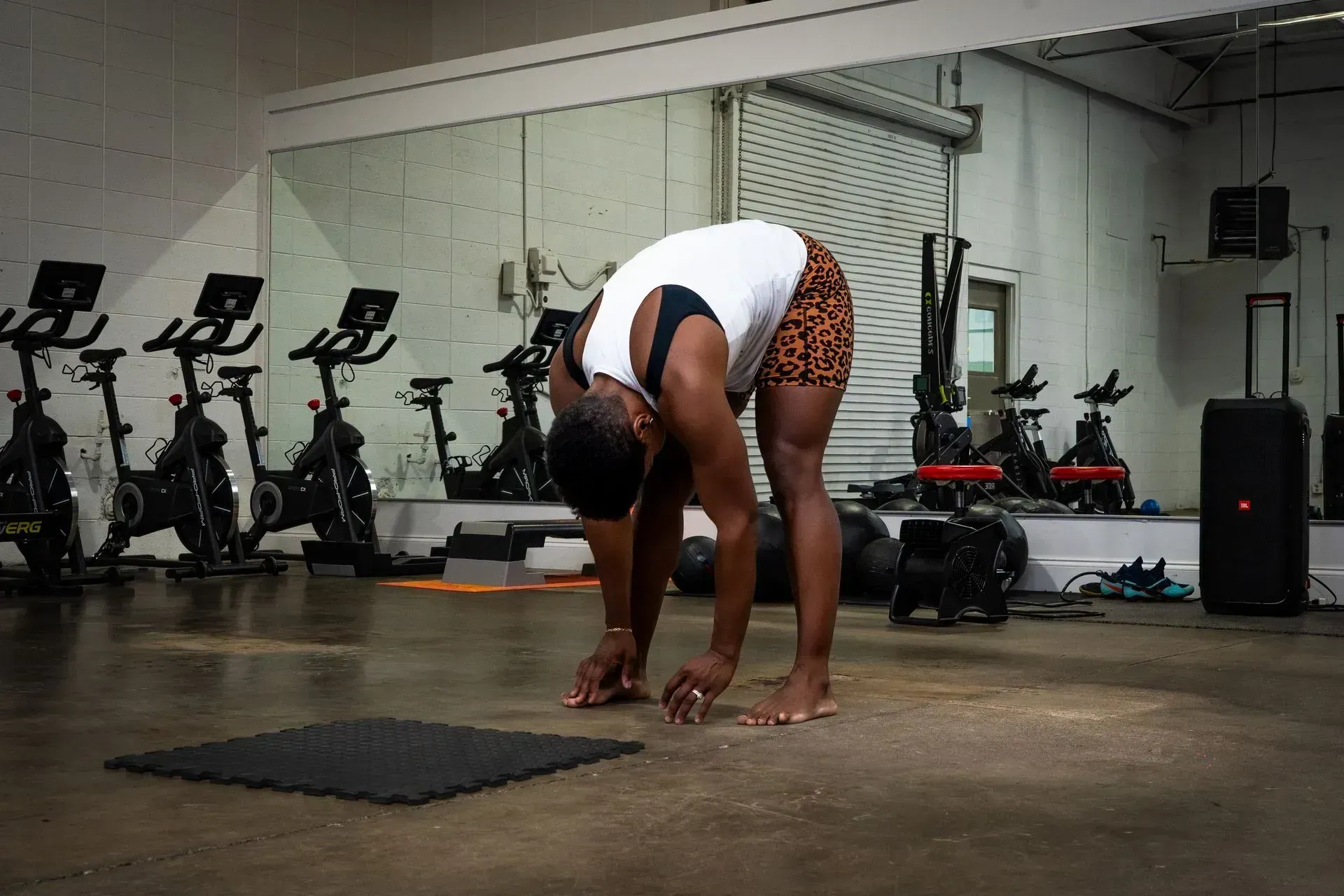 Person stretching in a gym, bending forward on a mat near stationary bikes and a punching bag.