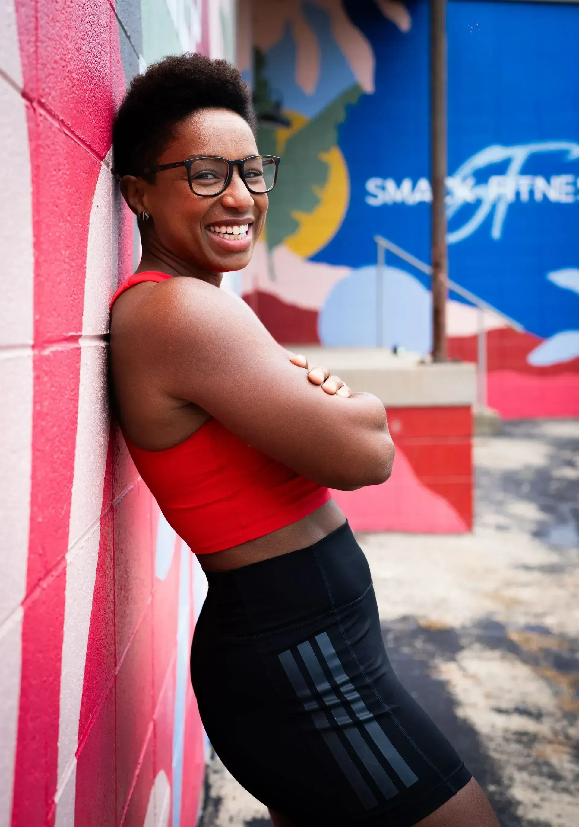 Smiling person in red top and black leggings leaning against a pink wall outdoors