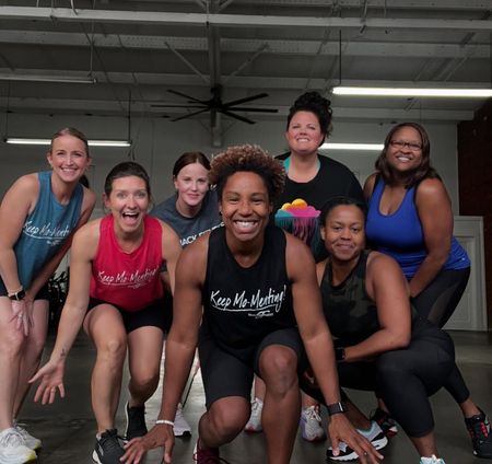 Group of seven smiling women posing in a gym, crouched and standing together in workout clothes.