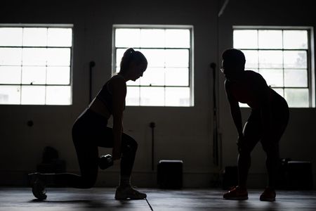 Two athletes in silhouette doing a kettlebell exercise in a gym with three bright windows.