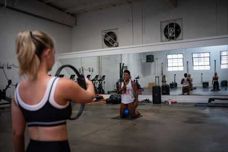 Person lifting weights in a gym, with a trainer seated in the background.