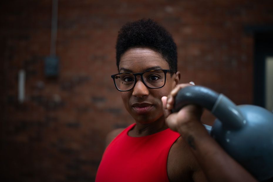 Person in glasses holding a kettlebell against a brick wall, wearing a red shirt.