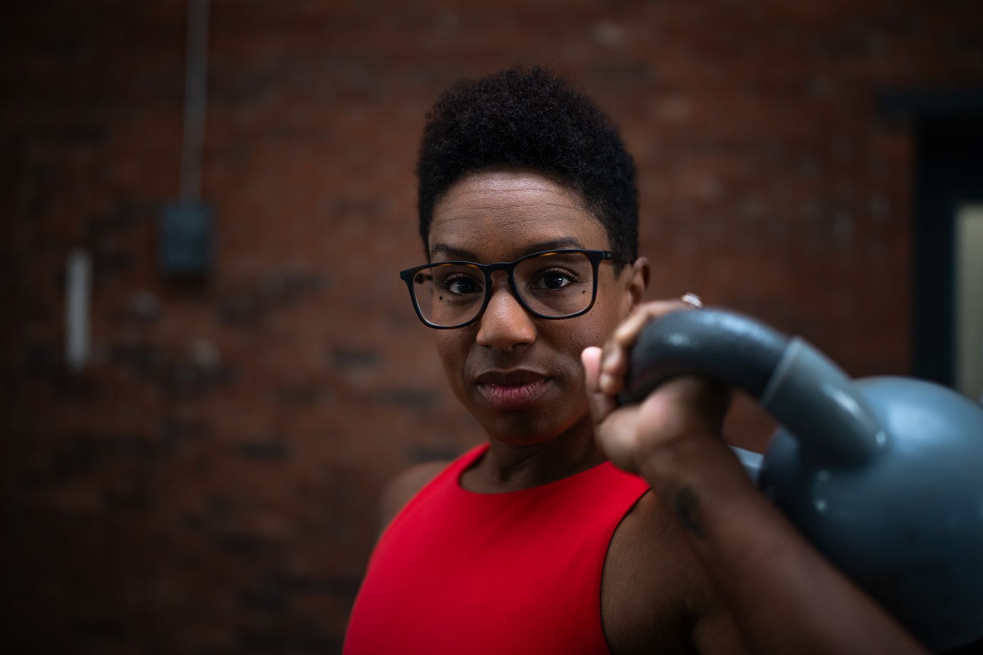 Person in glasses holding a kettlebell in a gym, wearing a red shirt against a brick wall.