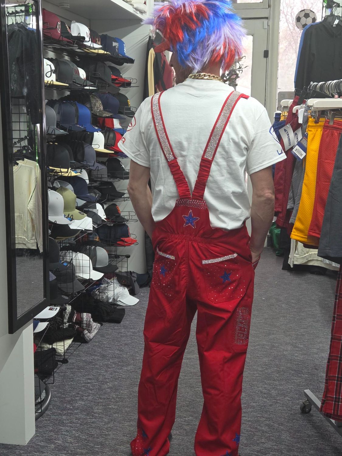 Person in red overalls and a blue-red wig standing in a clothing store aisle, seen from behind