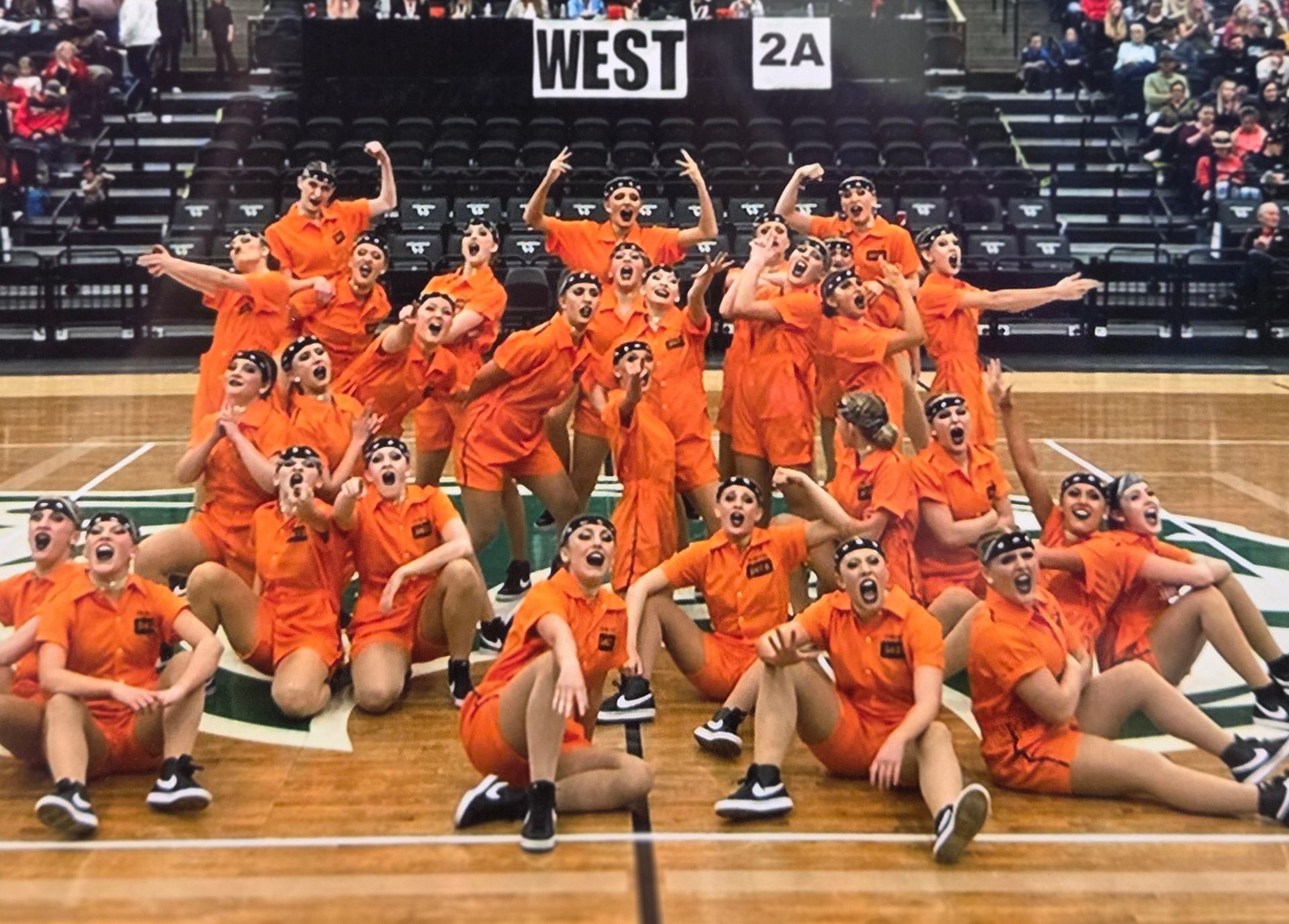Dance team in orange jumpsuits, poses on a basketball court. 