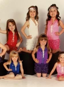 Six children posing in sparkly dance costumes against a plain backdrop