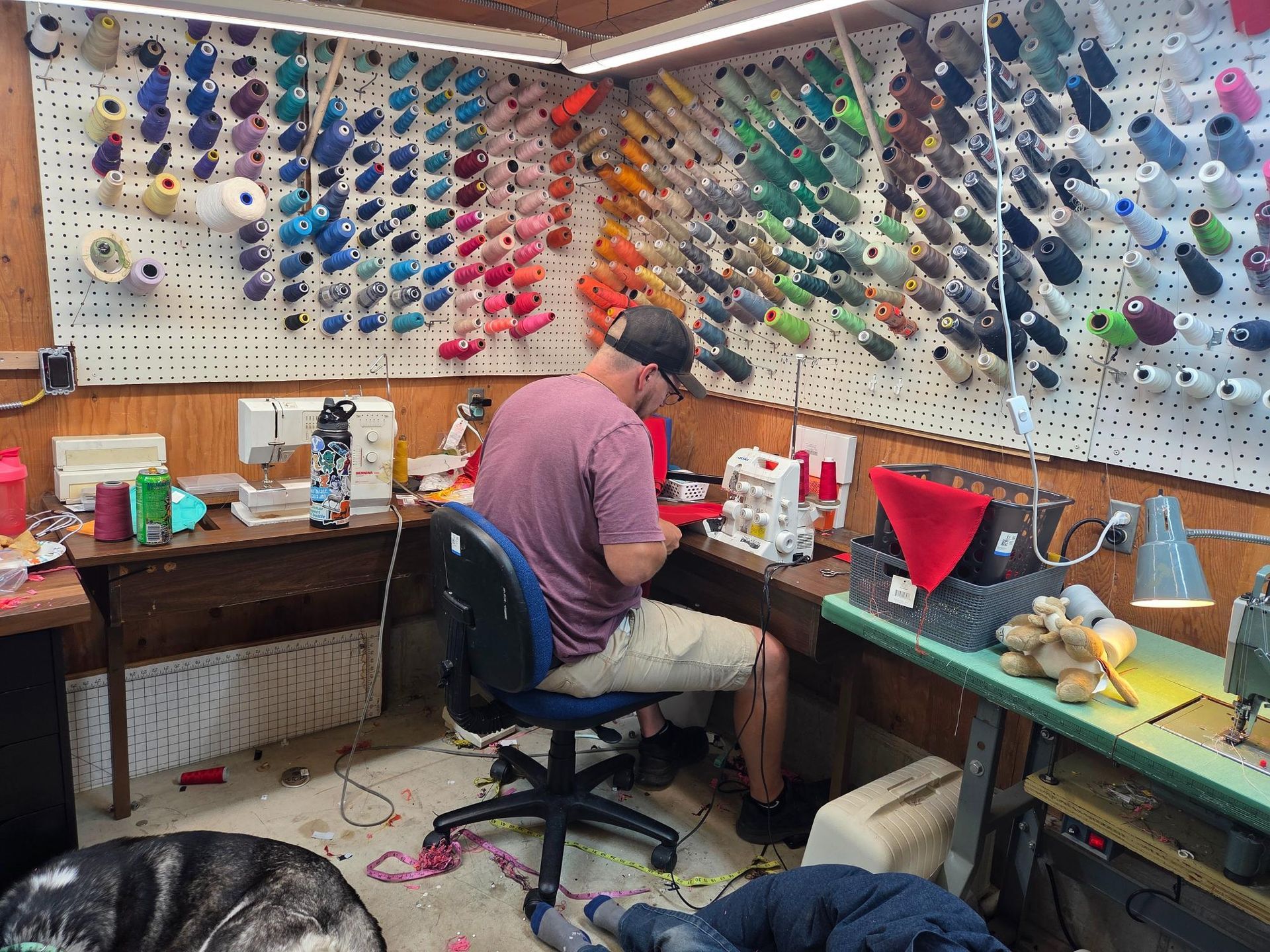 Man sewing at a cluttered workspace, with many colorful spools of thread on the wall. A dog lies on the floor.
