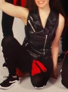 Two people kneeling, one in a black sleeveless outfit with red accents, against a plain background