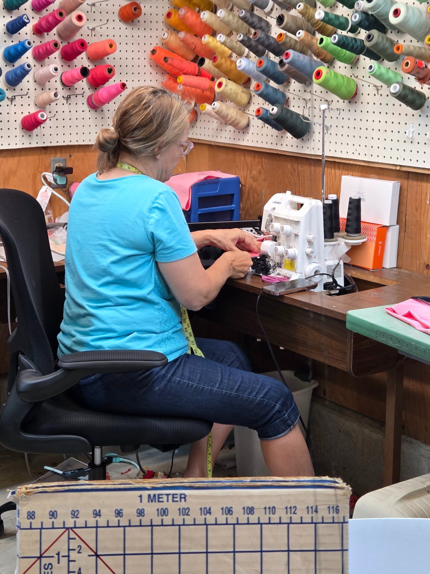 Woman in blue shirt sewing at a machine, surrounded by colorful spools of thread in a workshop.