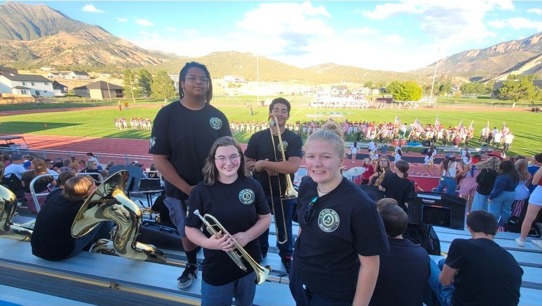 Band members posing with instruments on a field, mountains in the background, other band members further back.